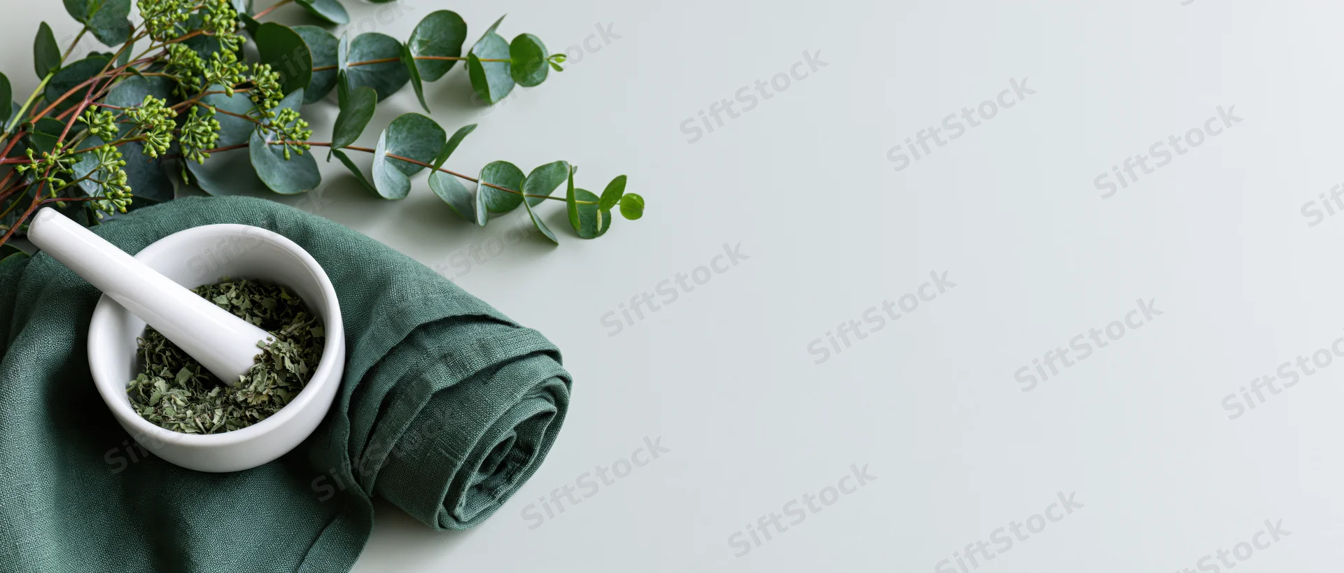 Eucalyptus branches and a white ceramic mortar with crushed herbs on a green cloth, arranged on a light surface with copyspace