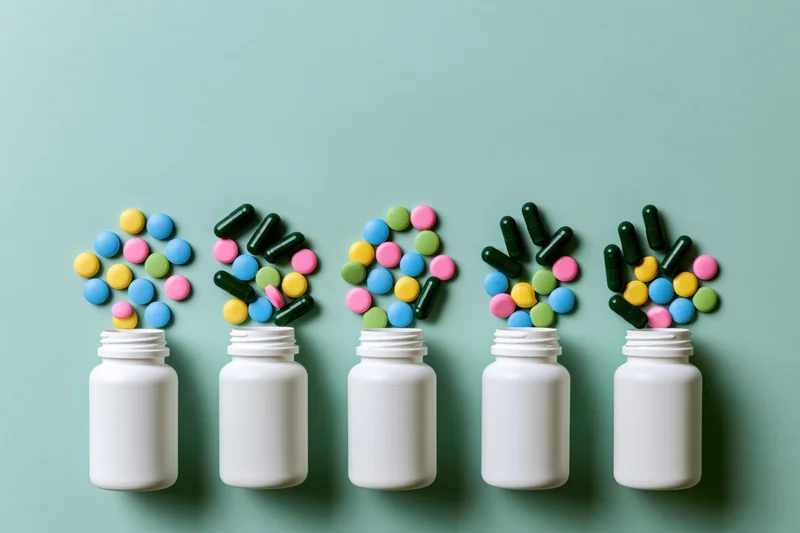 Five white pill bottles in a row with colorful pills spilling out on a mint green background