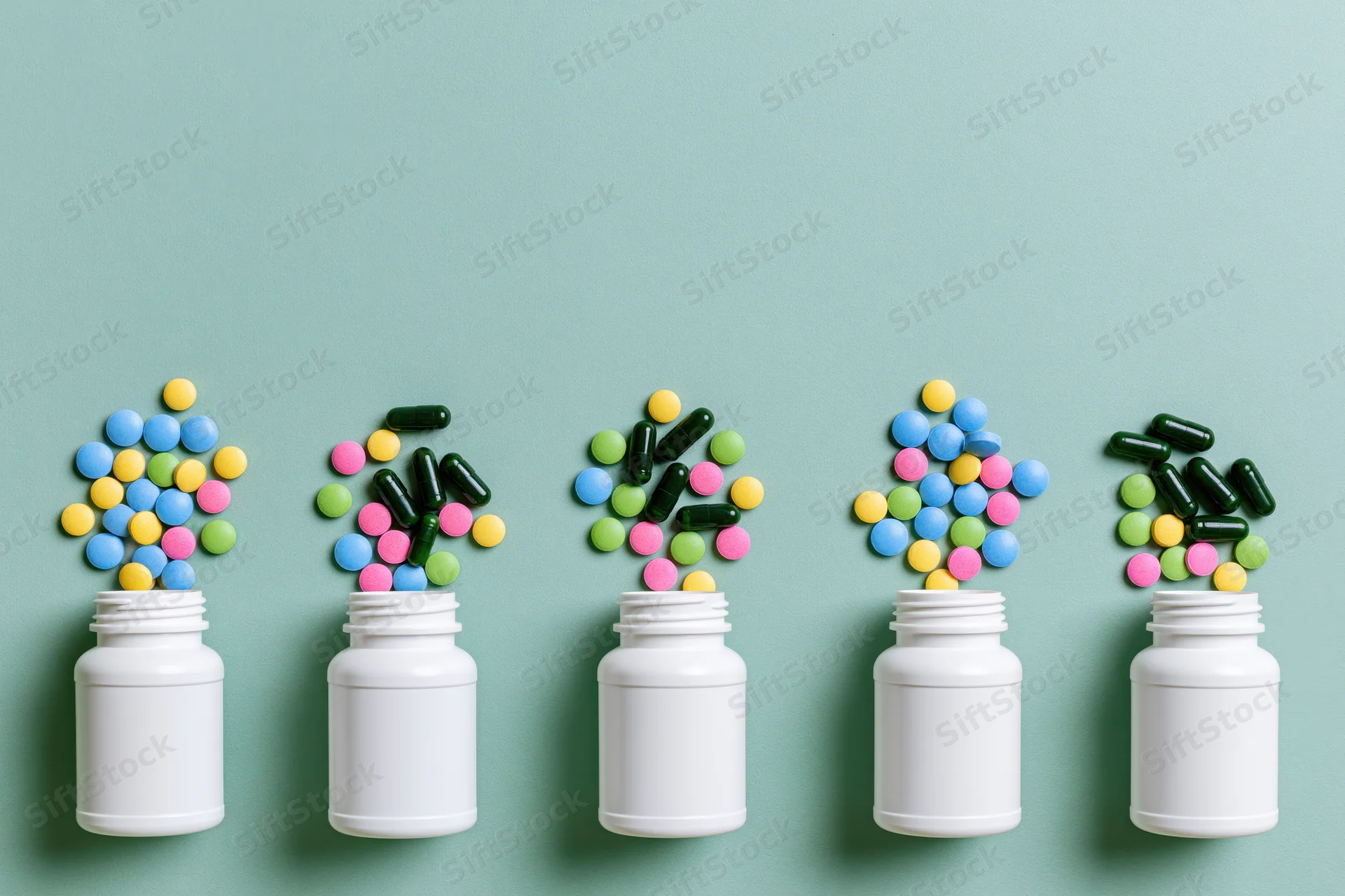 Five white pill bottles in a row with colorful capsules and tablets spilling out against a teal background