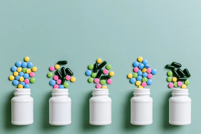 Five white pill bottles in a row with colorful capsules and tablets spilling out against a teal background