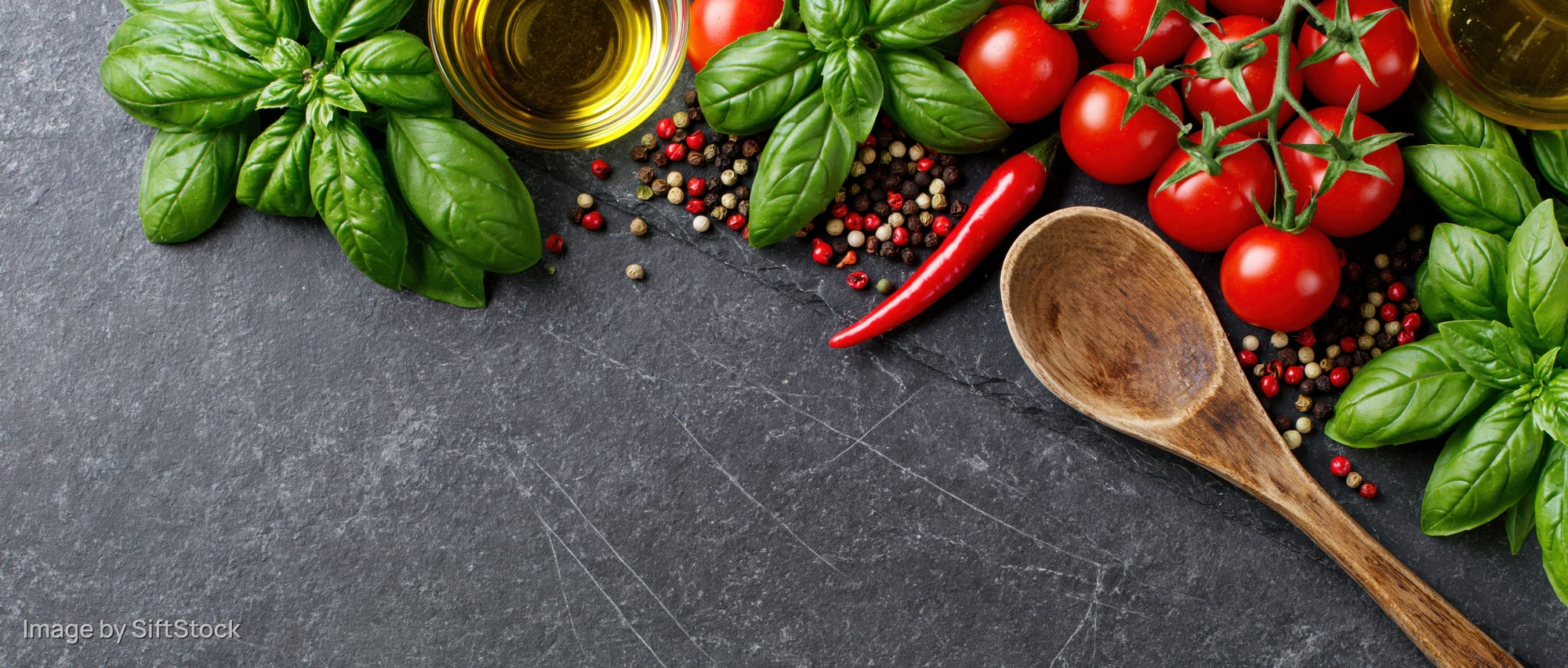 Fresh basil and cherry tomatoes with spices and olive oil arranged on dark slate surface with wooden spoon and copyspace