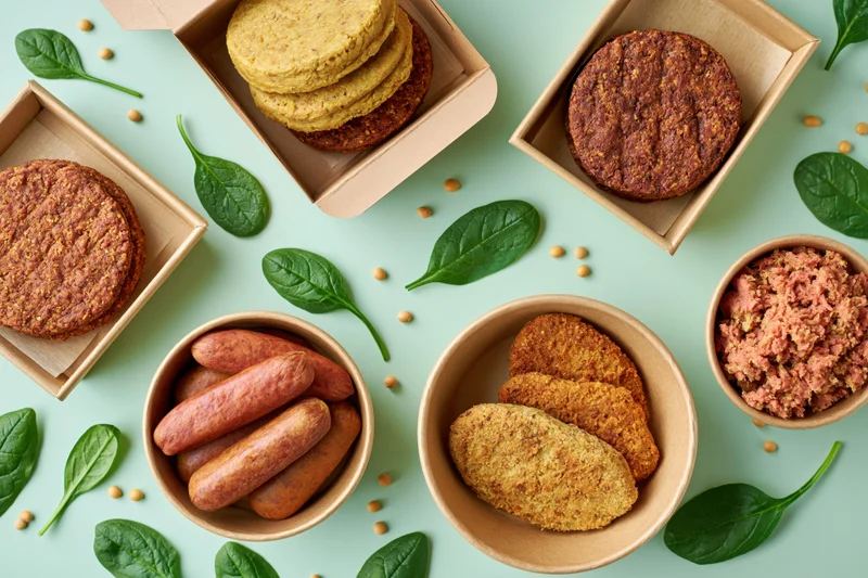 Assorted plant-based meat products in kraft paper boxes and bowls with spinach leaves on a light green surface