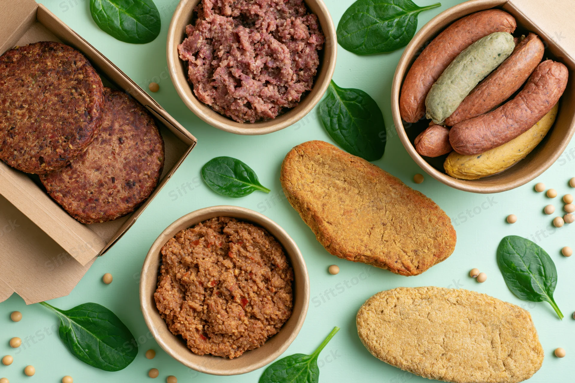 Assorted plant-based meat products arranged in kraft paper containers and bowls on a green surface with spinach and soybeans scattered around
