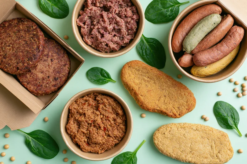 Assorted plant-based meat products arranged in kraft paper containers and bowls on a green surface with spinach and soybeans scattered around