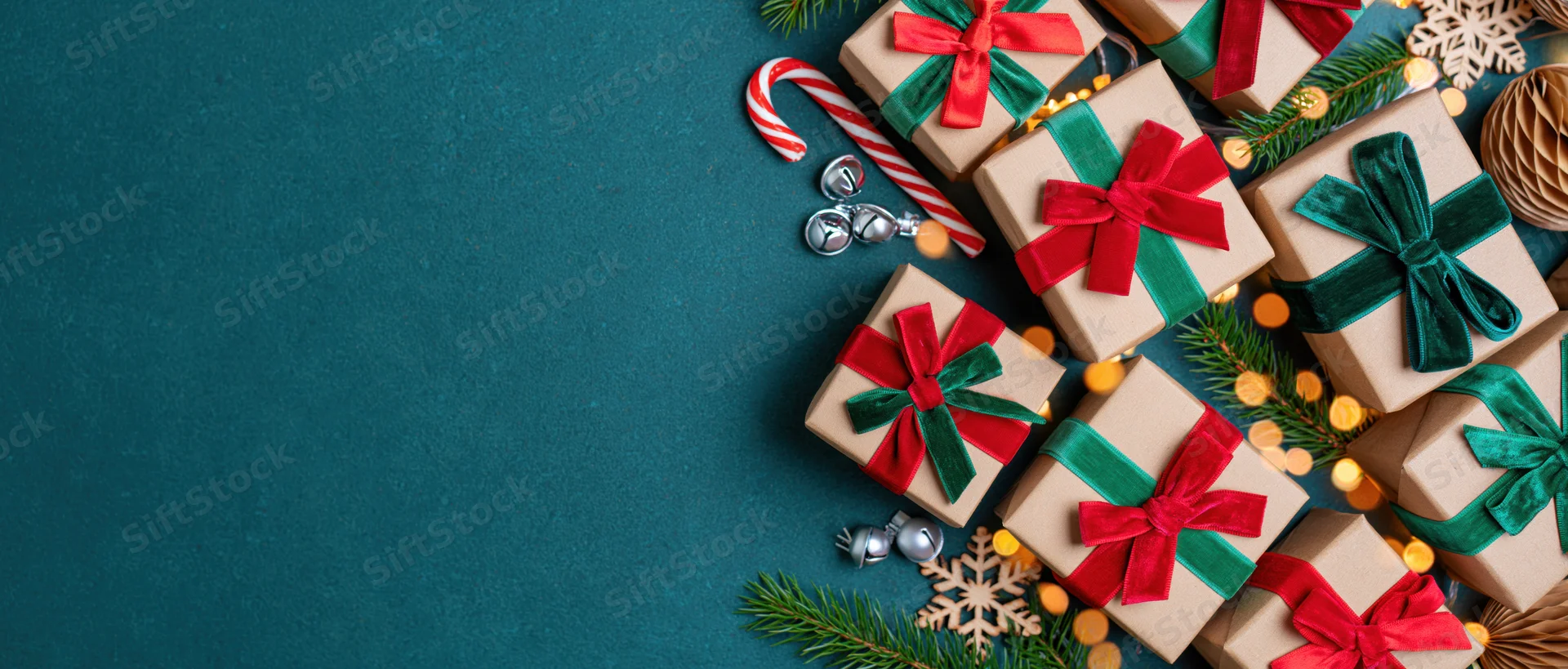 Kraft paper gift boxes with red and green velvet ribbons arranged with pine branches, candy canes, and ornaments on a teal surface