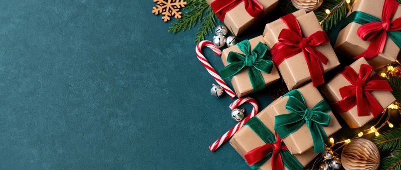 Kraft paper gift boxes with red and green bows, candy canes, pine branches and lights arranged on a teal textured surface in a flat lay composition with copyspace