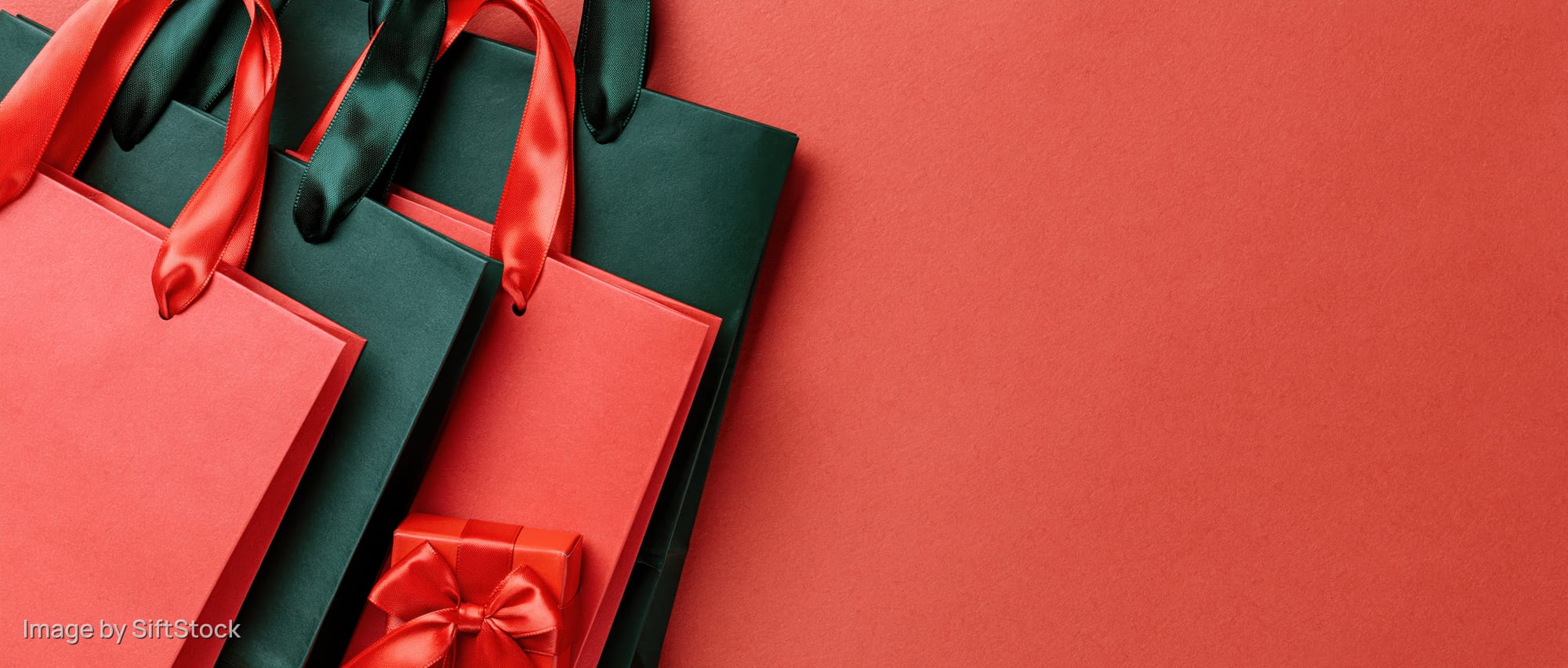 Red and dark green paper shopping bags with satin ribbons and a gift box arranged on a red surface