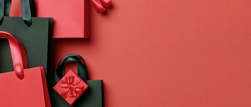 Red and dark green paper shopping bags with satin ribbons arranged in flat lay on red surface with copyspace