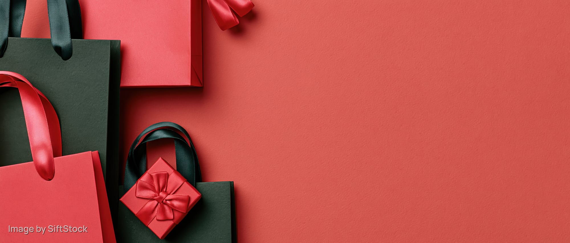 Red and dark green paper shopping bags with satin ribbons arranged in flat lay on red surface with copyspace
