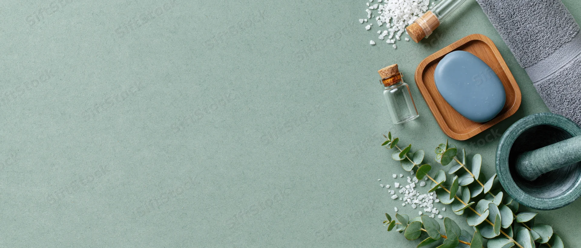 Spa and skincare items arranged on right side of green surface with eucalyptus and salt