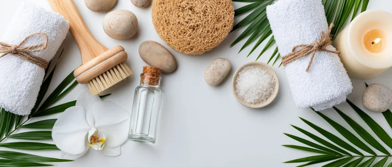 Spa wellness accessories arranged in flat lay on white surface with towels, brush, loofah, stones, candle, salt, orchid and palm leaves