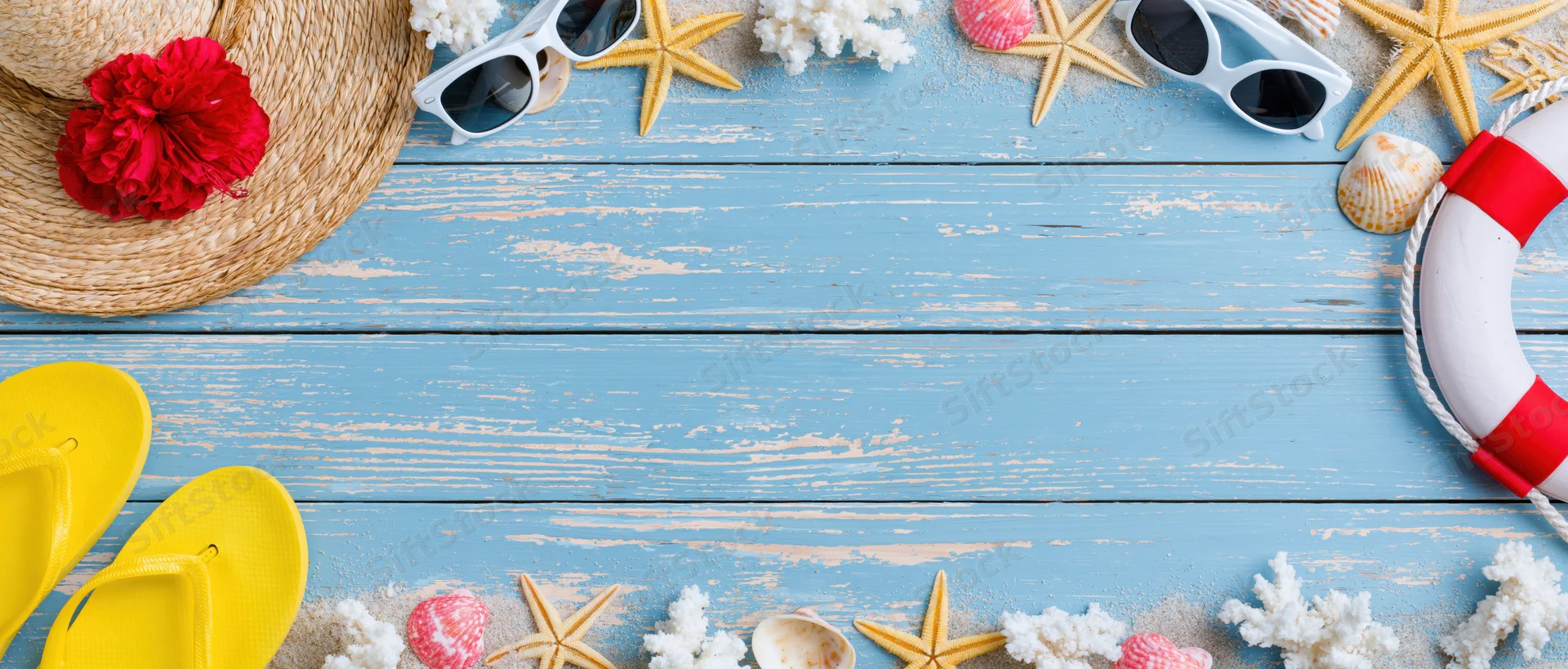 Summer beach accessories arranged along top and bottom edges of blue wooden surface with copyspace in center
