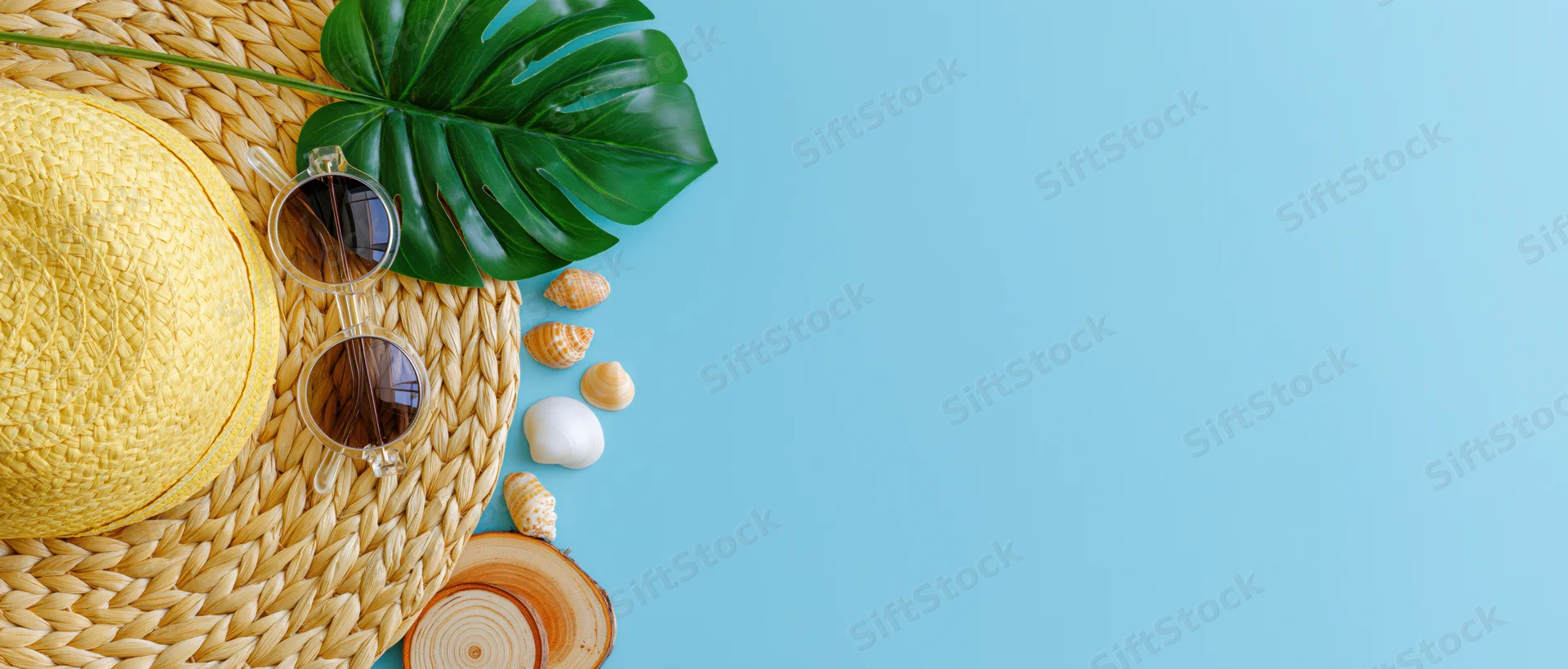 Summer beach accessories arranged on woven mat with monstera leaf and shells on blue background