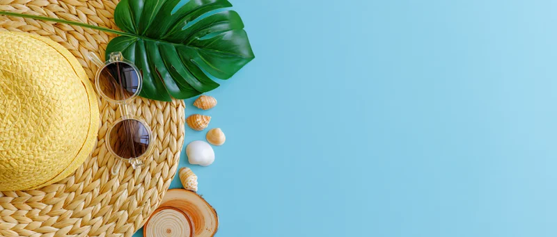 Summer beach accessories arranged on woven mat with monstera leaf and shells on blue background