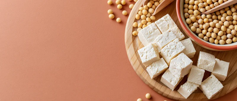 Cubed tofu and soybeans arranged on a wooden board with a ceramic bowl on a solid background