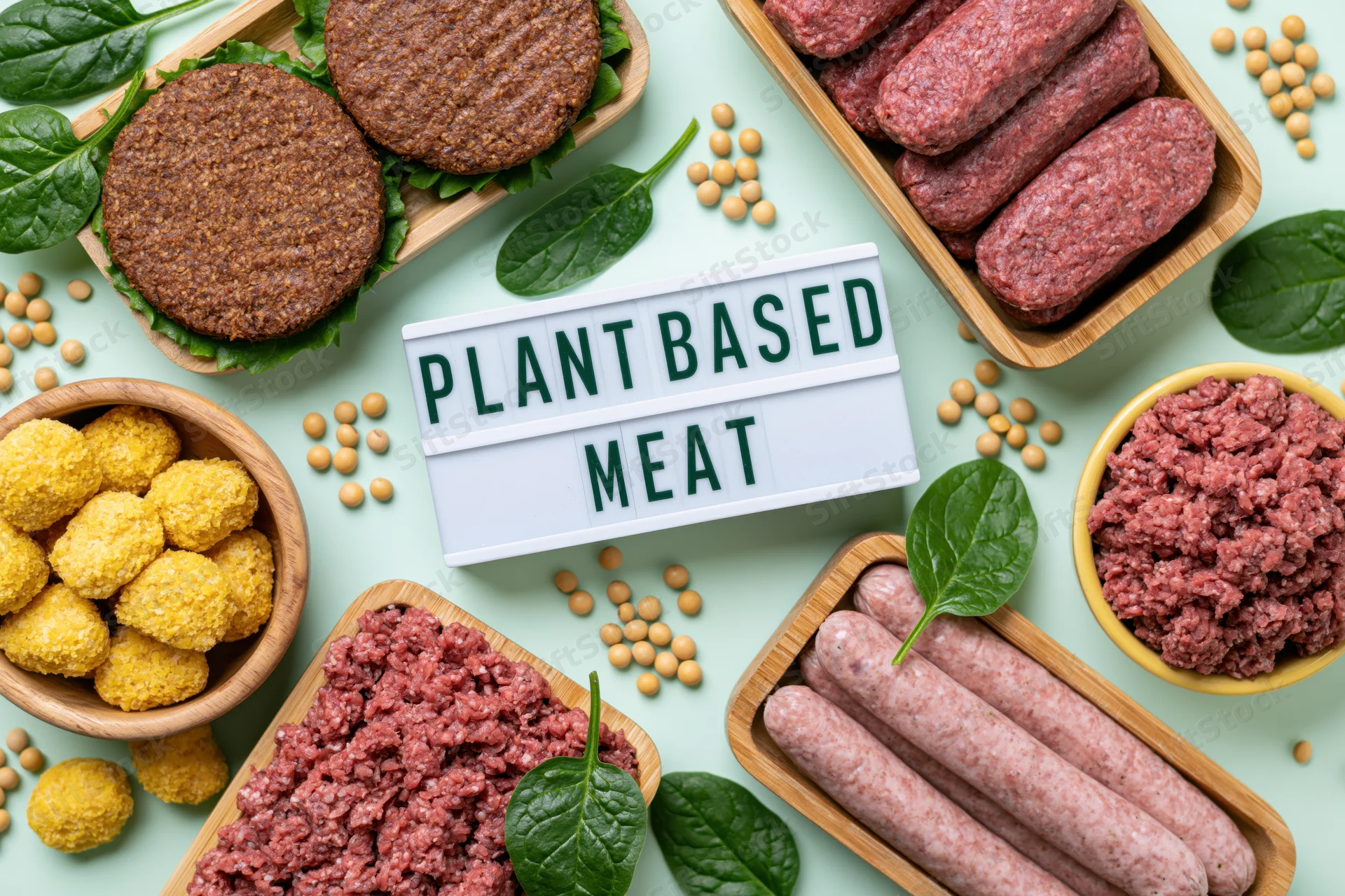 Plant-based meat products arranged in wooden trays and bowls with soybeans and spinach on a light green surface