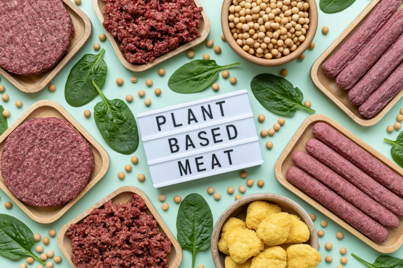 Plant-based meat products arranged in wooden trays and bowls with soybeans and spinach on a light blue surface, centered around a lightbox sign reading "PLANT BASED MEAT