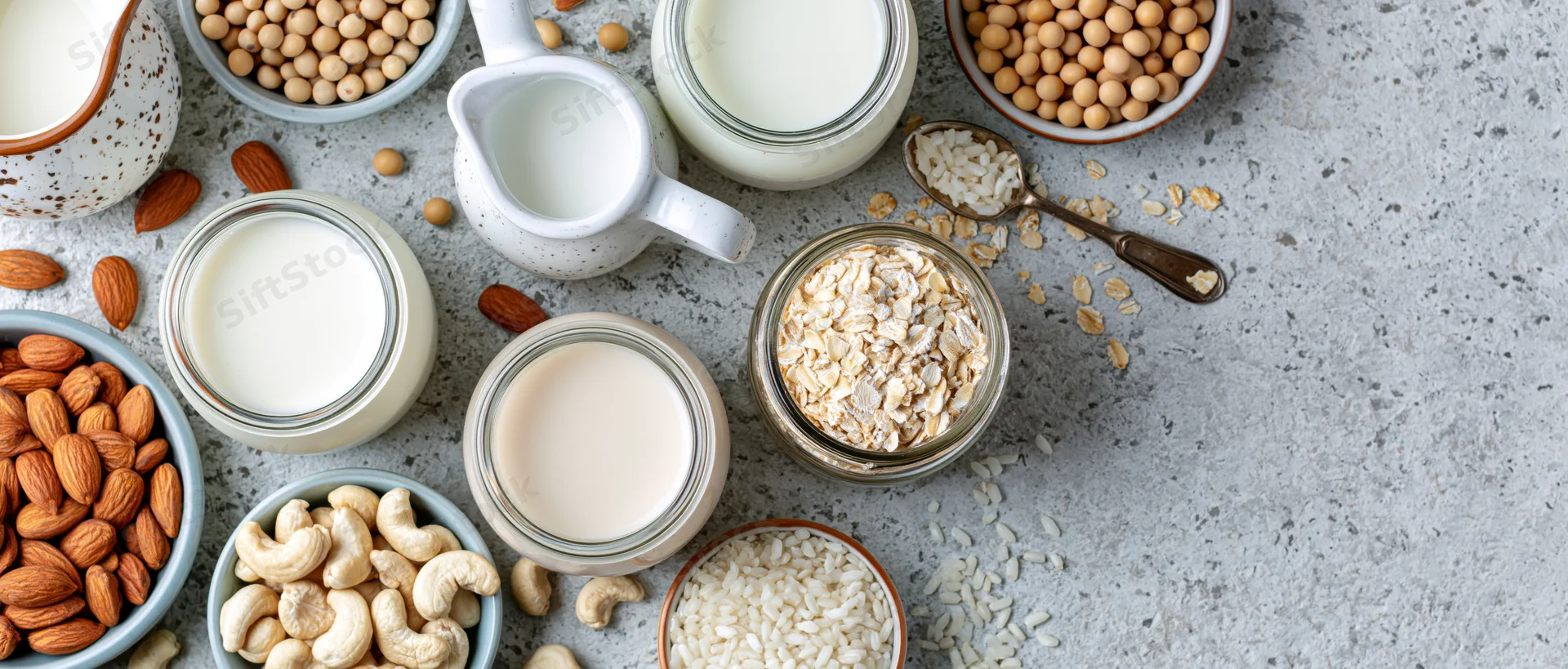 Various plant-based milk alternatives in glass jars and ceramic pitchers arranged on a gray countertop with nuts and grains