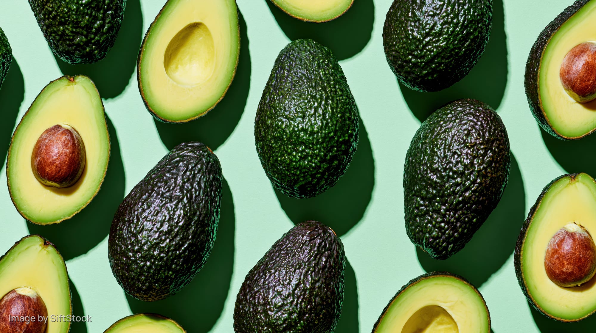 Whole and halved avocados arranged in a scattered flat lay on a green background