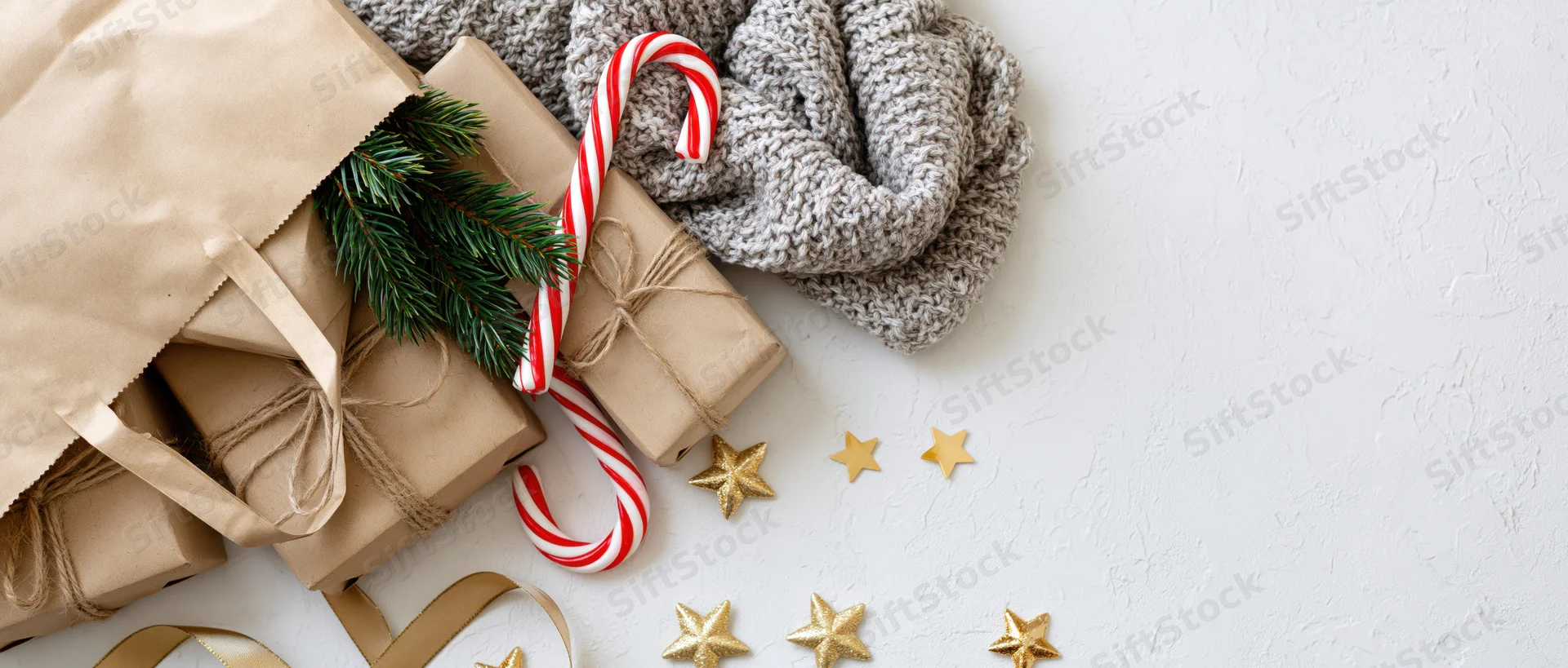 Kraft paper bag with wrapped gifts, candy cane, pine branch and knit scarf on white textured surface