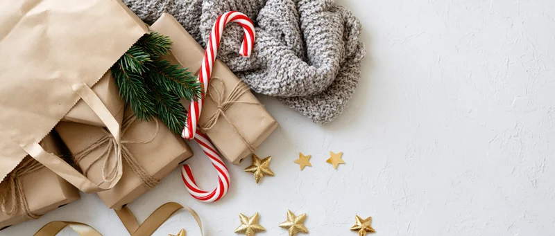 Kraft paper bag with wrapped gifts, candy cane, pine branch and knit scarf on white textured surface