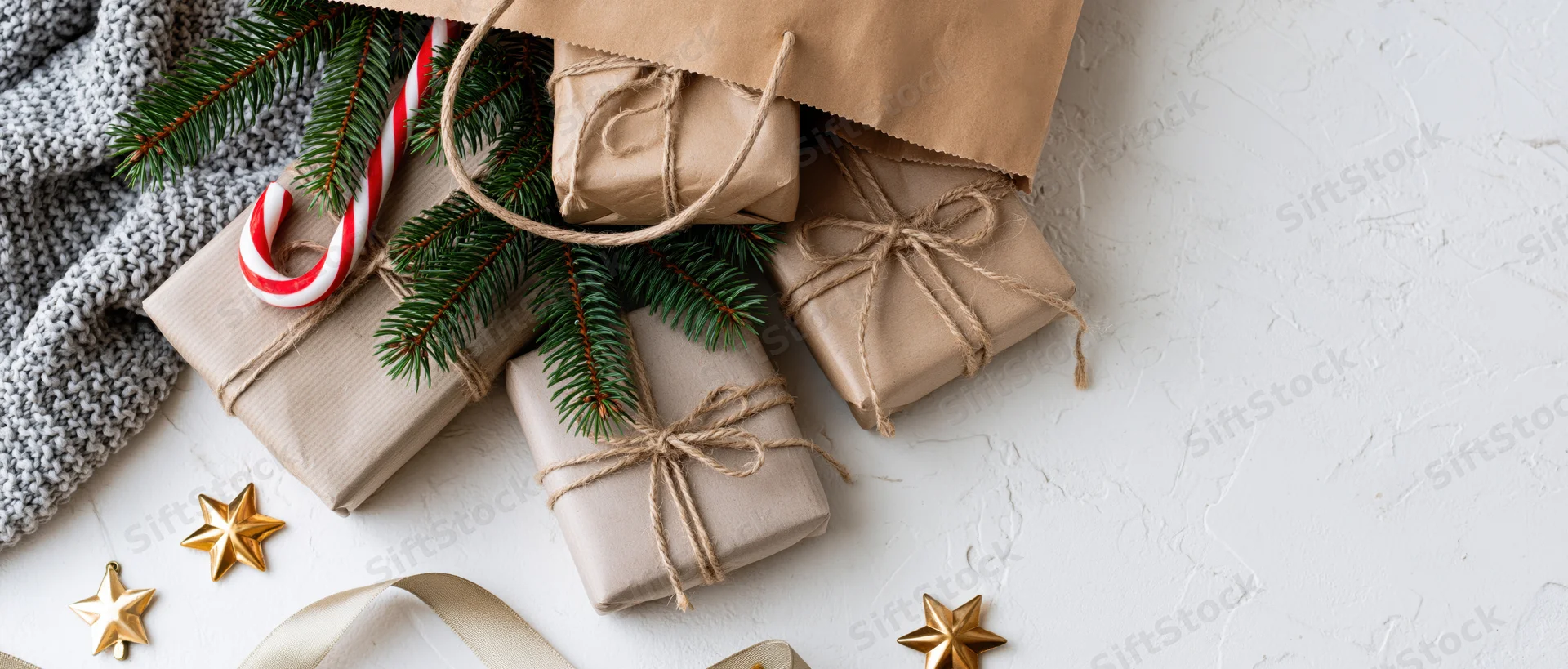 Kraft paper bag tipped over with wrapped gifts, evergreen sprigs, candy cane, and golden stars on white textured surface
