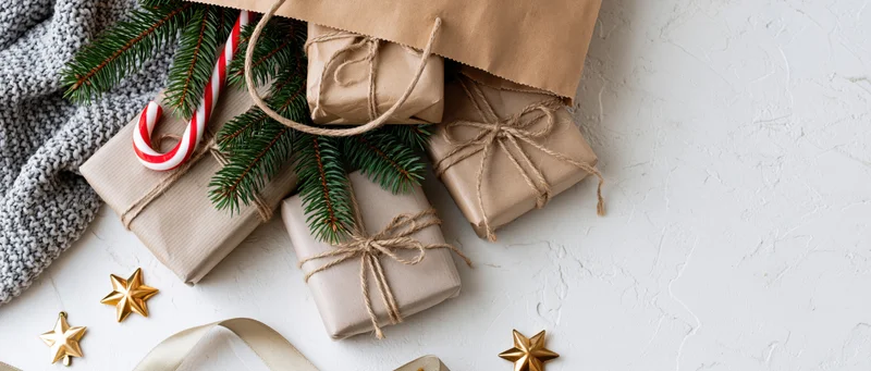 Kraft paper bag tipped over with wrapped gifts, evergreen sprigs, candy cane, and golden stars on white textured surface