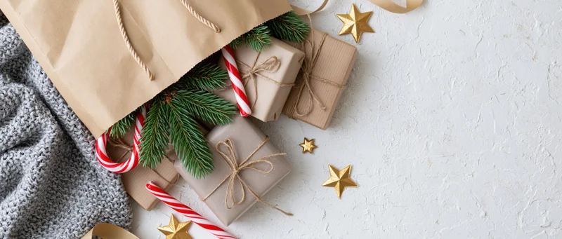Kraft paper bag spilling wrapped gifts, candy canes and pine sprigs on a white textured surface with gold stars and copyspace