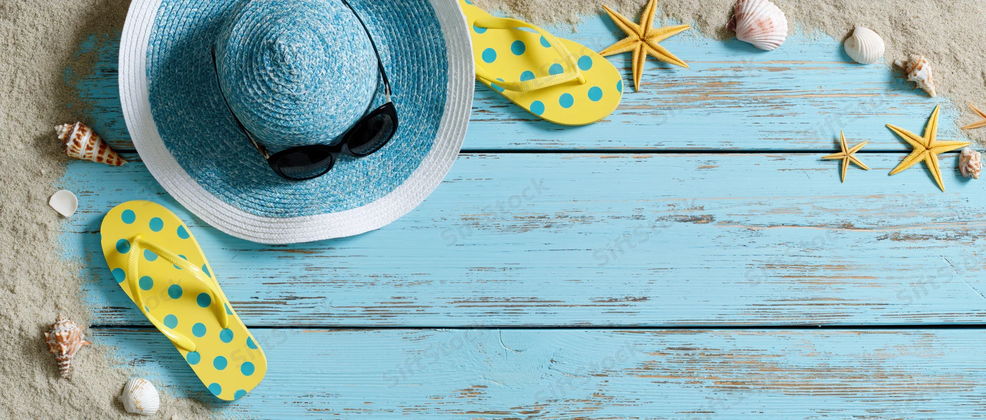 Summer beach accessories arranged on a blue wooden deck with sand and shells, overhead flat lay view