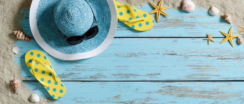 Summer beach accessories arranged on a blue wooden deck with sand and shells, overhead flat lay view