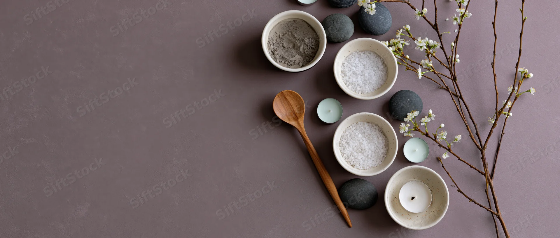 Spa arrangement with bowls of salt, powder, candles, stones and blossoms on muted mauve surface