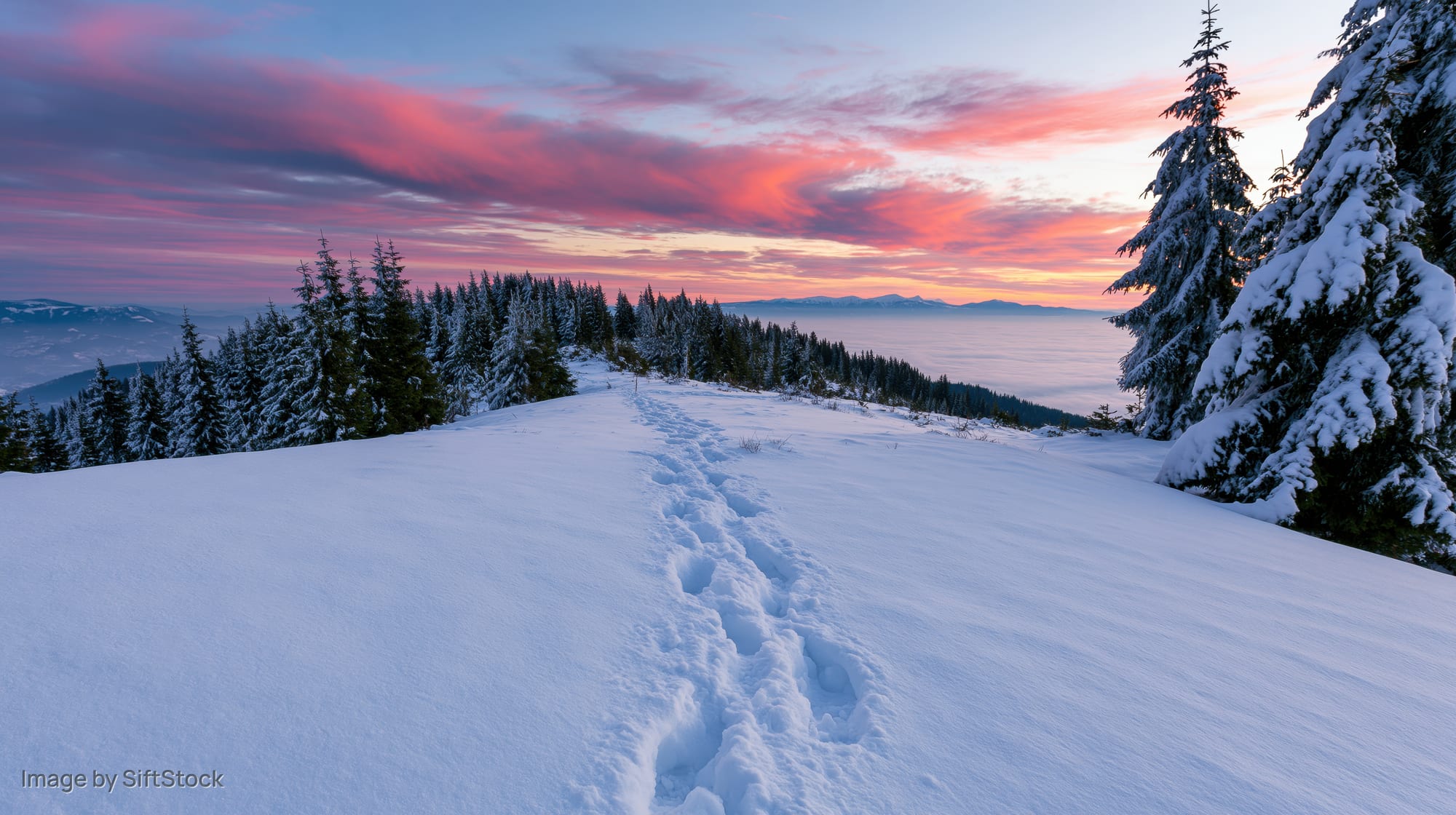 Footprints in deep snow leading across a snowy mountain ridge toward a colorful sunset sky