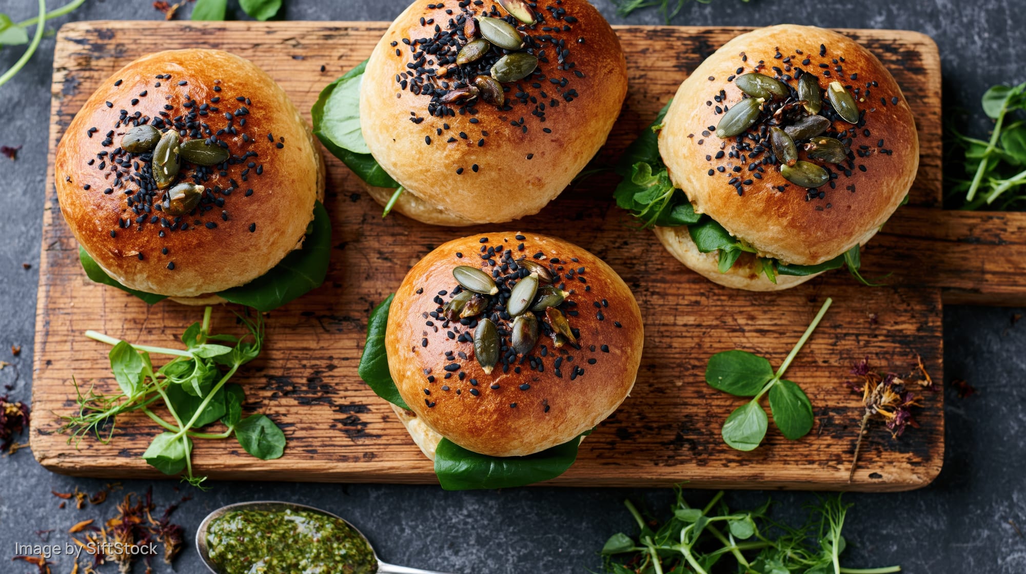 Four golden brioche sliders topped with black sesame and pumpkin seeds on a wooden cutting board with fresh greens and pesto spoon nearby