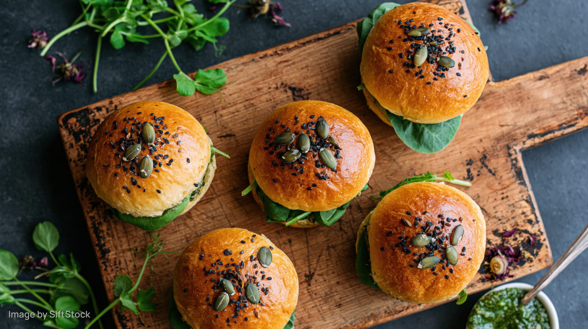 Five golden brioche sliders topped with black sesame and pumpkin seeds on a wooden cutting board with greens and herb sauce