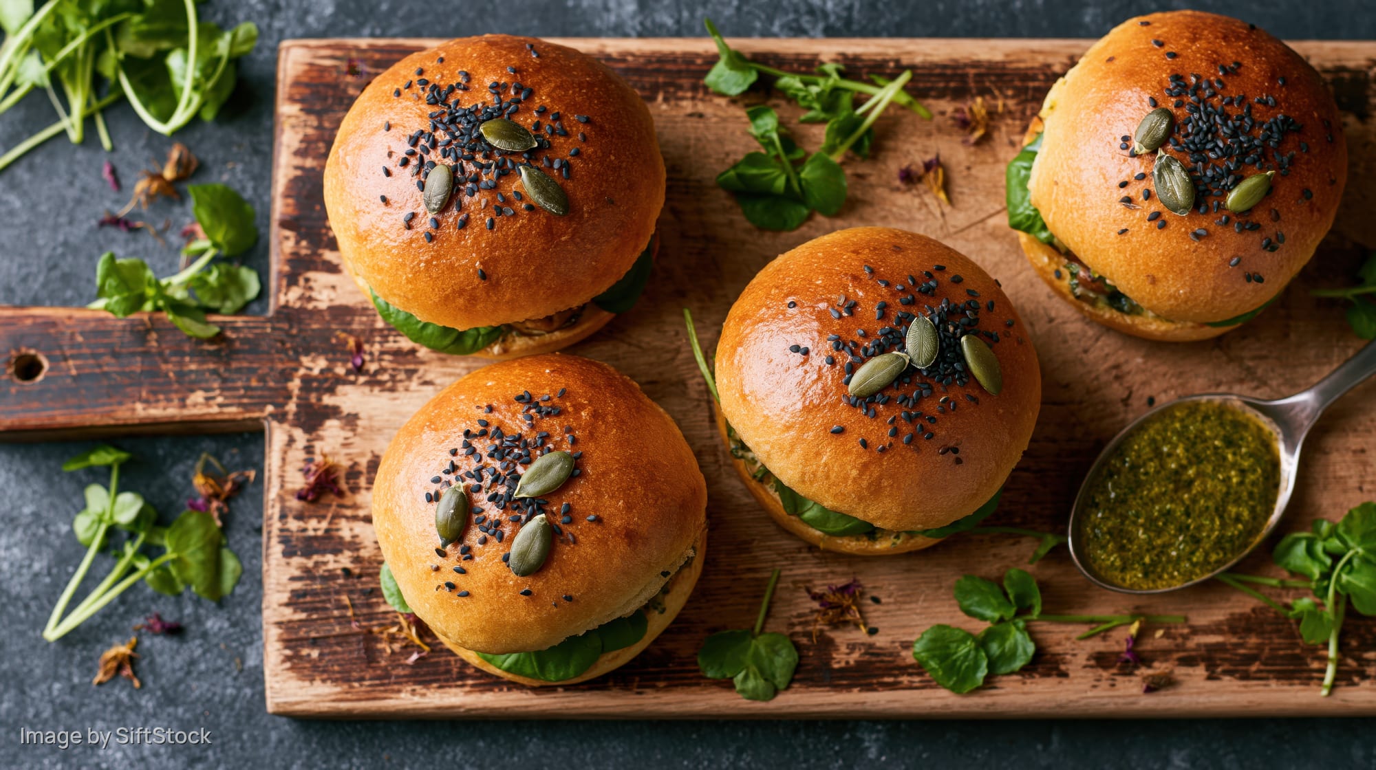 Four golden brioche sliders topped with black sesame and pumpkin seeds on a wooden cutting board with pesto and microgreens