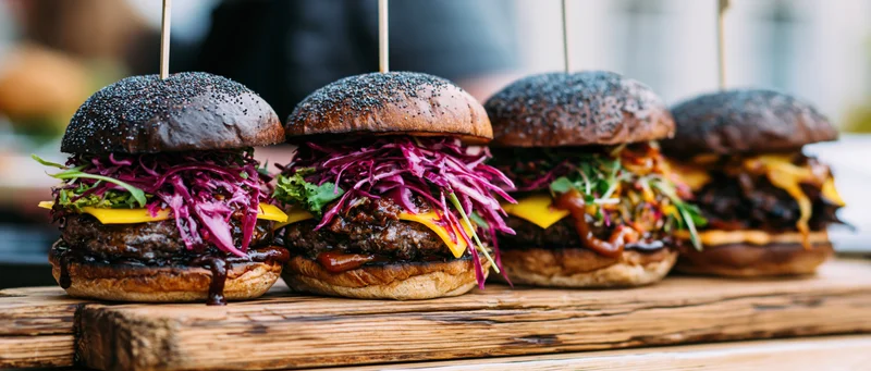 Four gourmet burgers with dark poppy-seed brioche buns lined on a wooden board