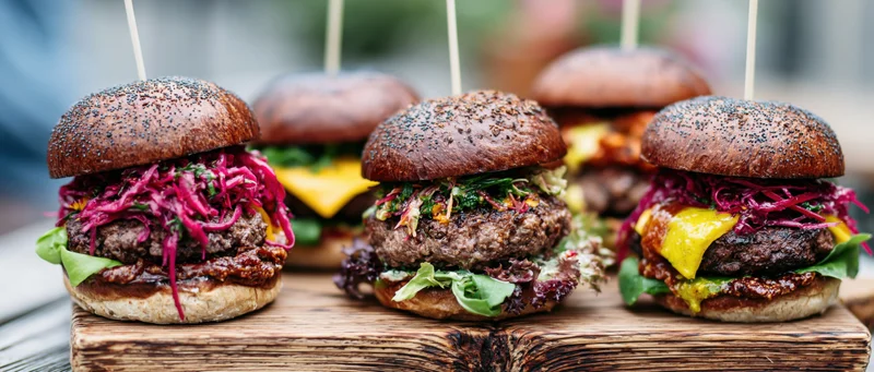Four gourmet burgers with dark poppy-seed brioche buns and colorful toppings arranged on a wooden board