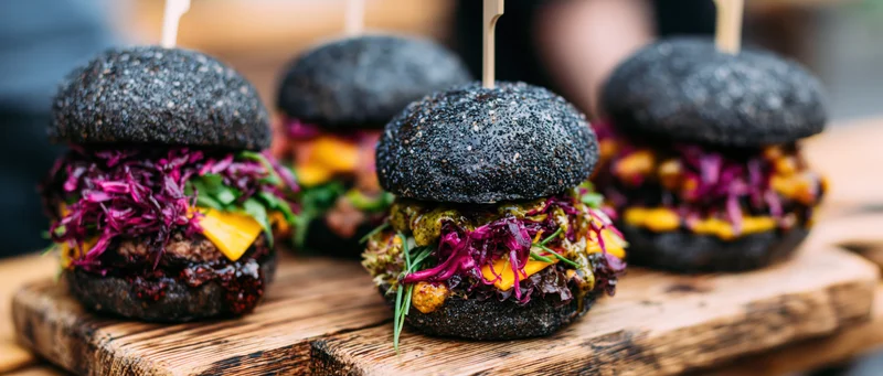 Four gourmet burgers with black poppyseed brioche buns and colorful toppings arranged on a wooden board
