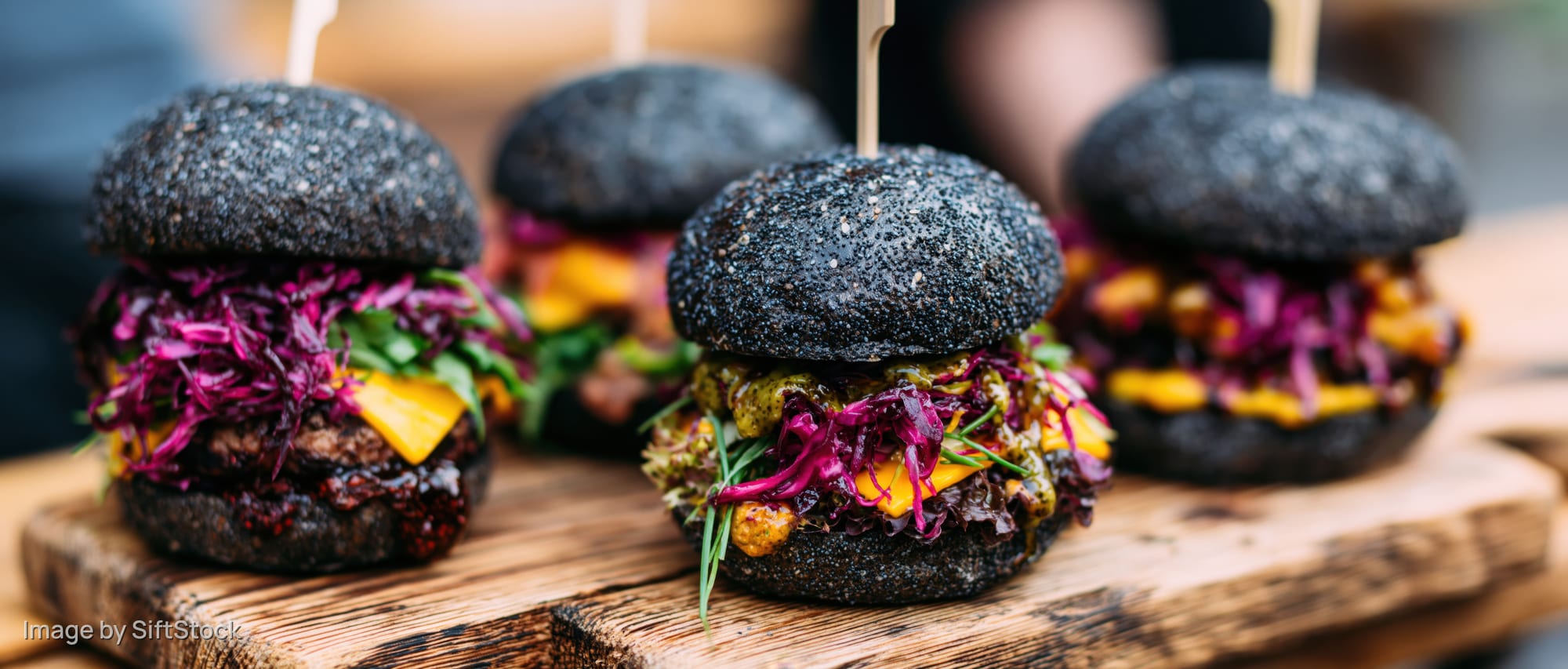 Four gourmet burgers with black poppyseed brioche buns and colorful toppings arranged on a wooden board