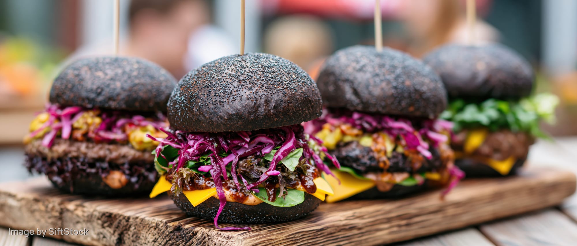 Four gourmet burgers with dark poppy-seed brioche buns lined on a wooden board