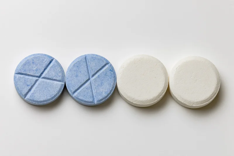 Four round pharmaceutical tablets arranged in a row on a white surface, two blue scored and two white smooth