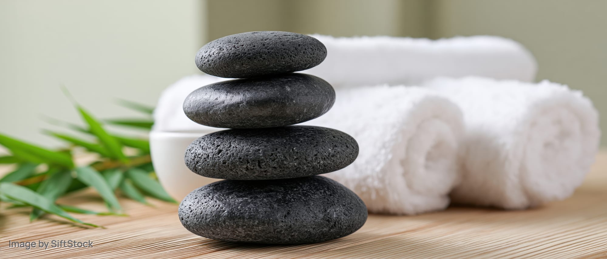 Four dark basalt massage stones stacked on a bamboo mat beside white rolled towels and green leaves