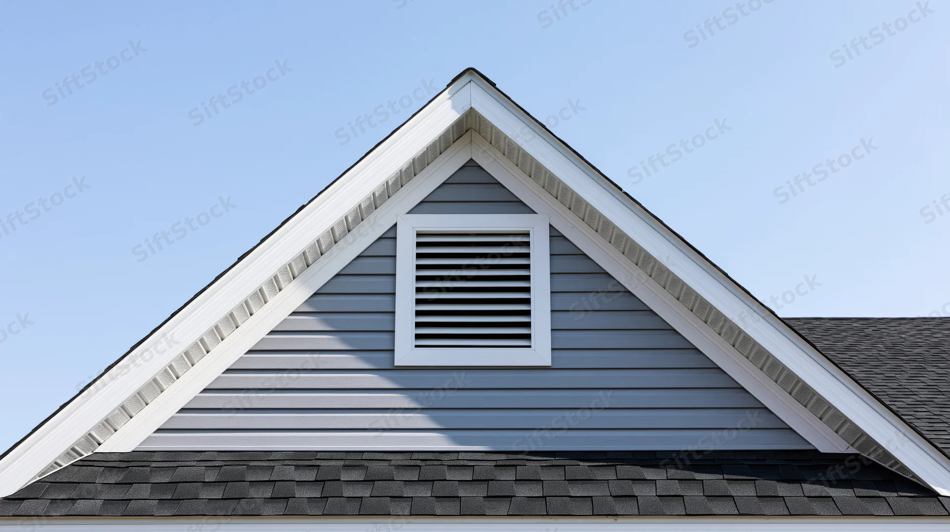 Gable end of a residential house with louvered attic vent and gray siding under a clear blue sky