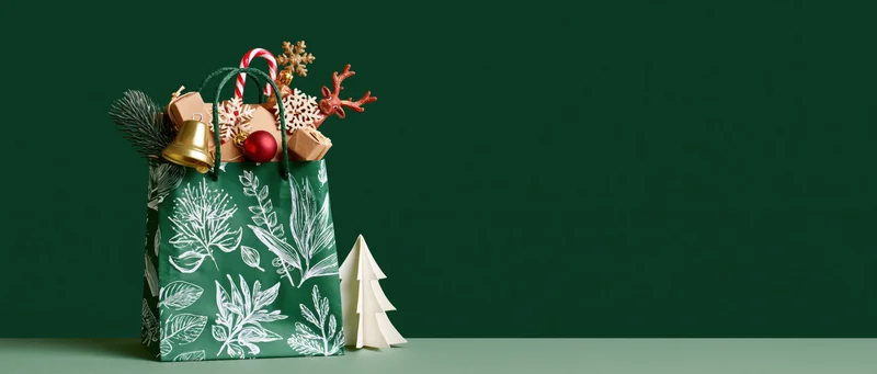 Green gift bag with white botanical print and christmas ornaments on dark green surface
