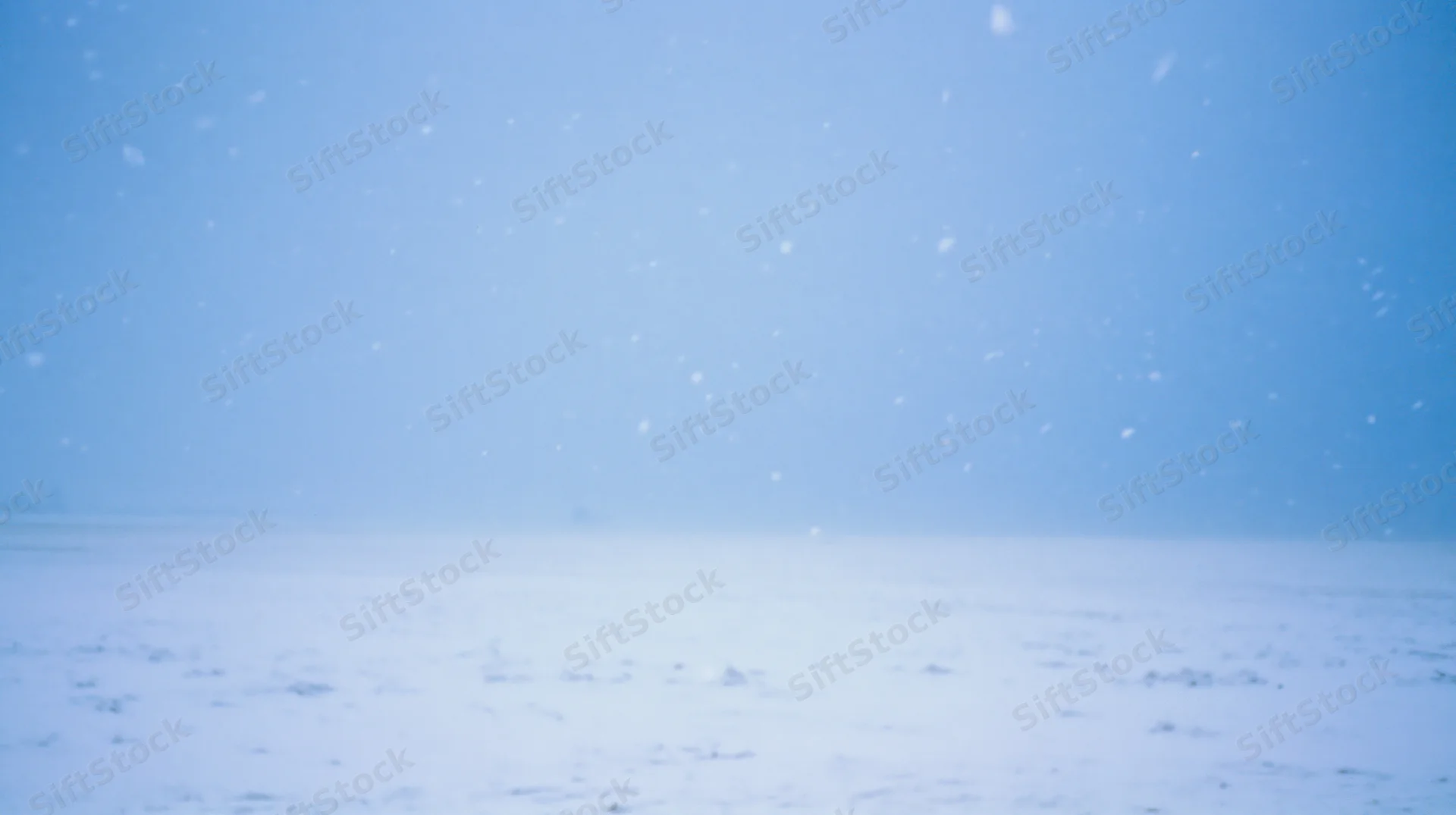 Snow falling over a flat snowy field under a pale blue sky