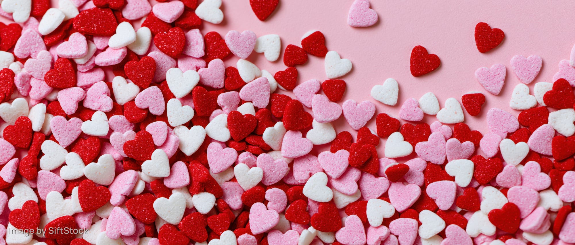 Heart-shaped sugar sprinkles scattered across a pink surface in a flat lay arrangement