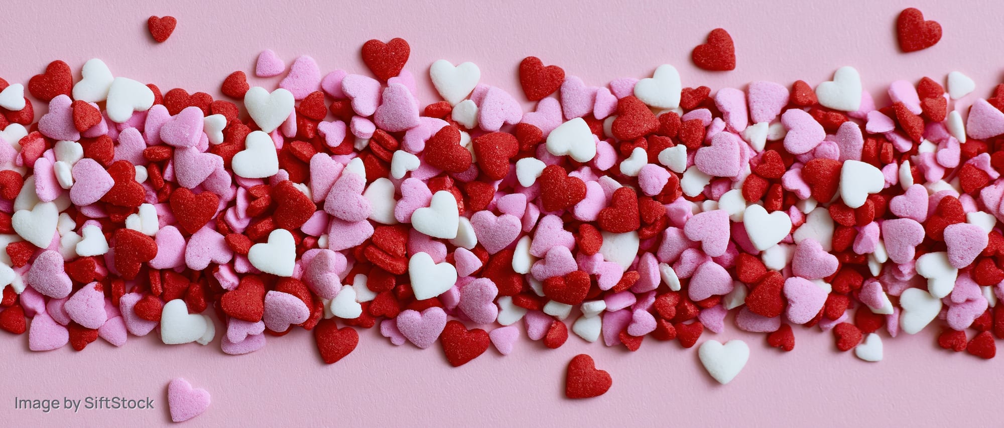 Heart-shaped sugar sprinkles scattered across a pink surface in a flat lay arrangement