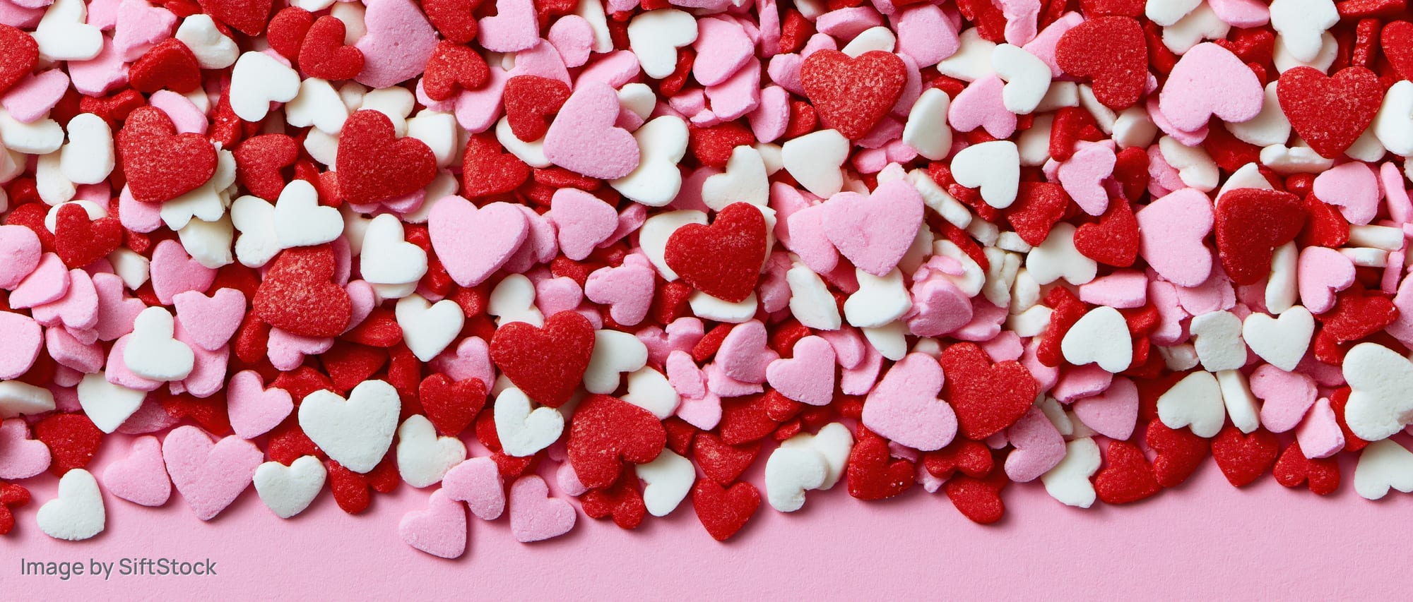 Heart-shaped sugar sprinkles scattered across a pink surface with copyspace at the bottom