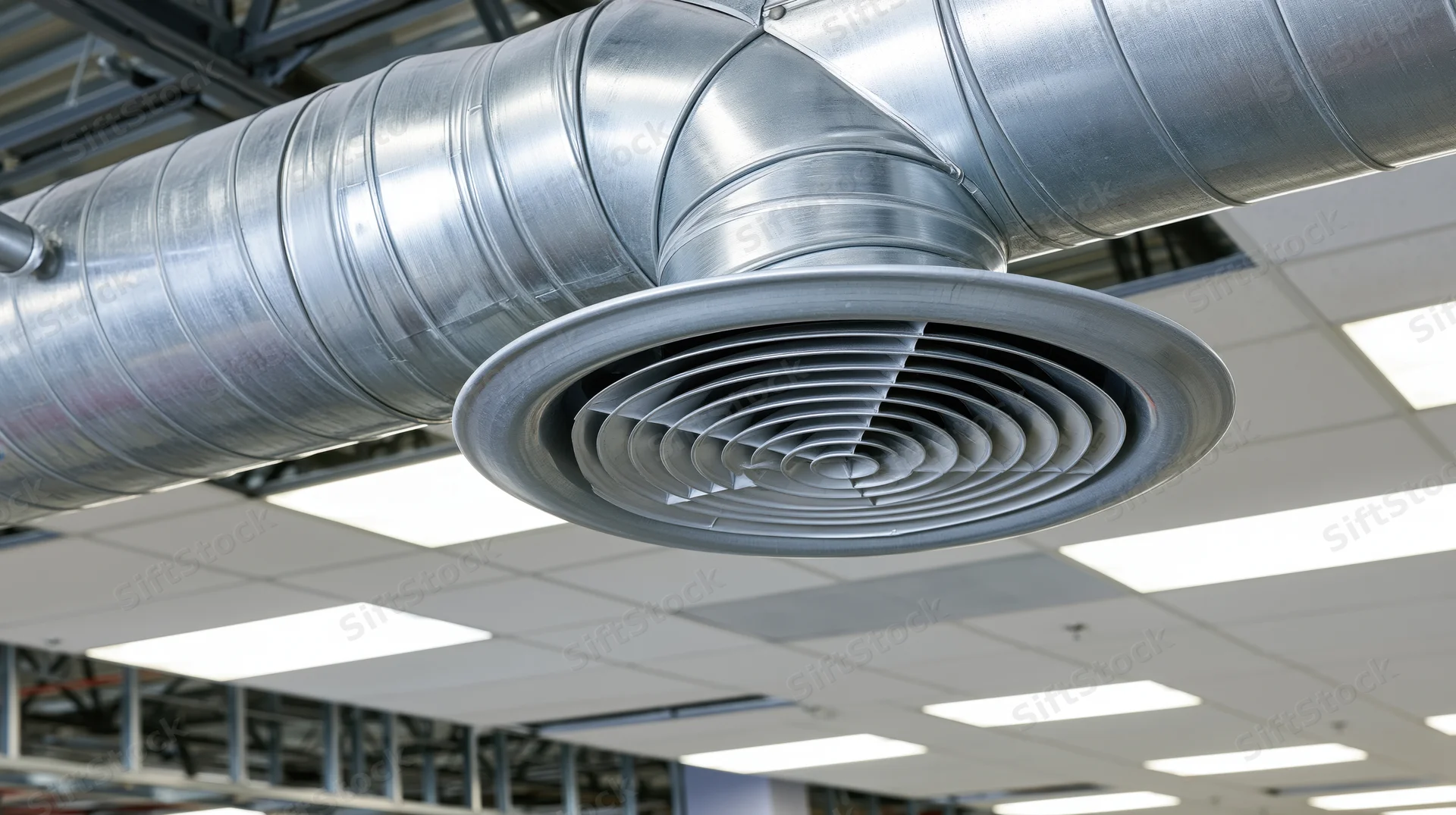 Large metallic hvac duct with spiral grille mounted on industrial ceiling under fluorescent lighting