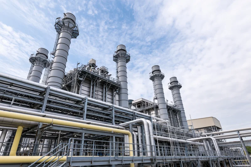 Industrial gas turbine power plant with multiple tall metal exhaust stacks and interconnected piping under a partly cloudy sky